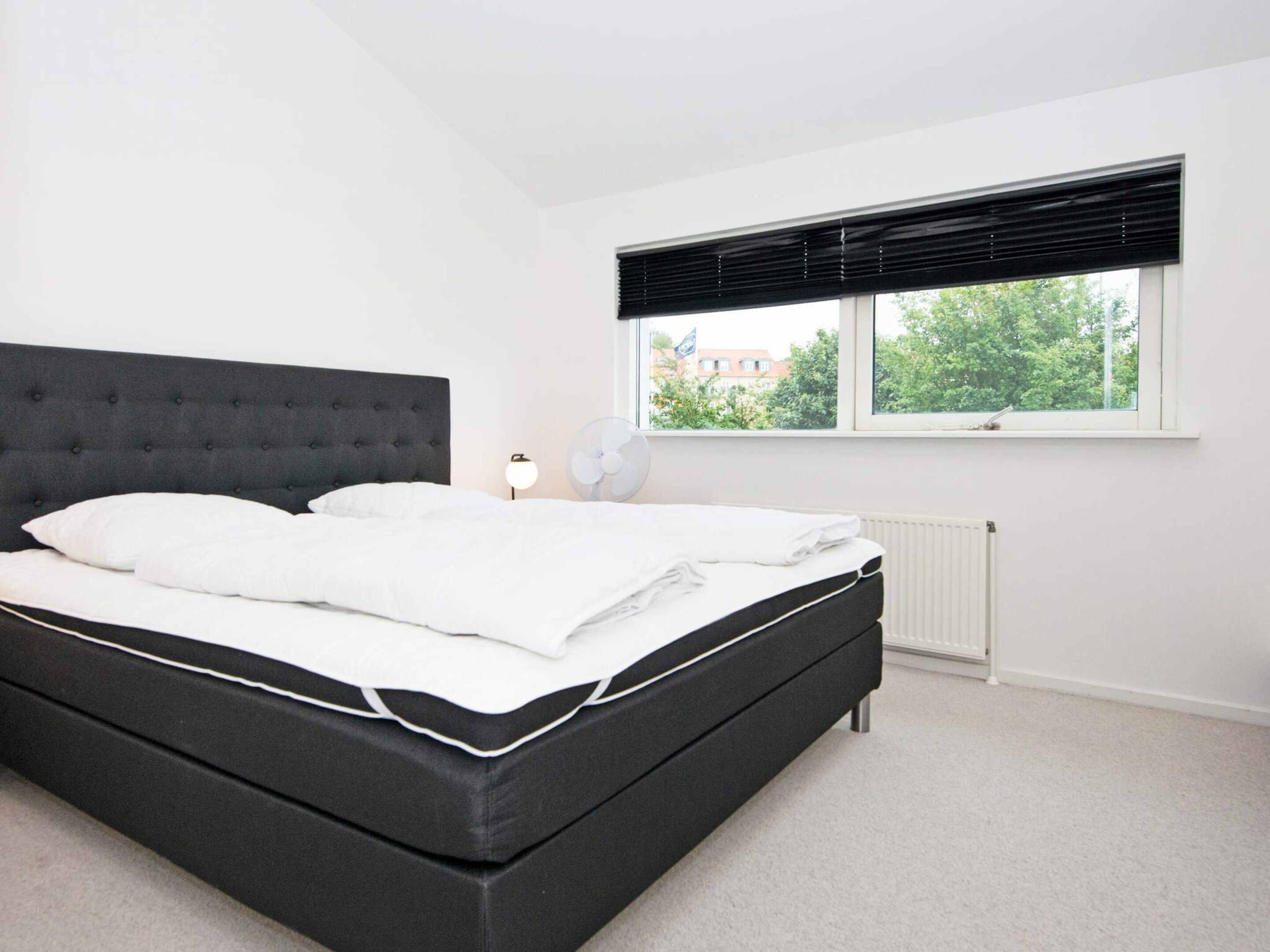 A modern bedroom with a large bed, a window with blinds, a radiator, and a bedside table with a lamp.