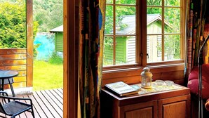 A wooden deck with a table and chairs, a window with a view of a green house and trees, and a small cabinet with a lantern and books.