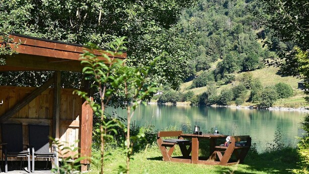 A rustic wooden cabin with a dining table and chairs, overlooking a calm lake surrounded by lush greenery.