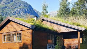 A wooden cabin with a sloped roof, a small porch, and a window overlooking a grassy area with mountains in the background.