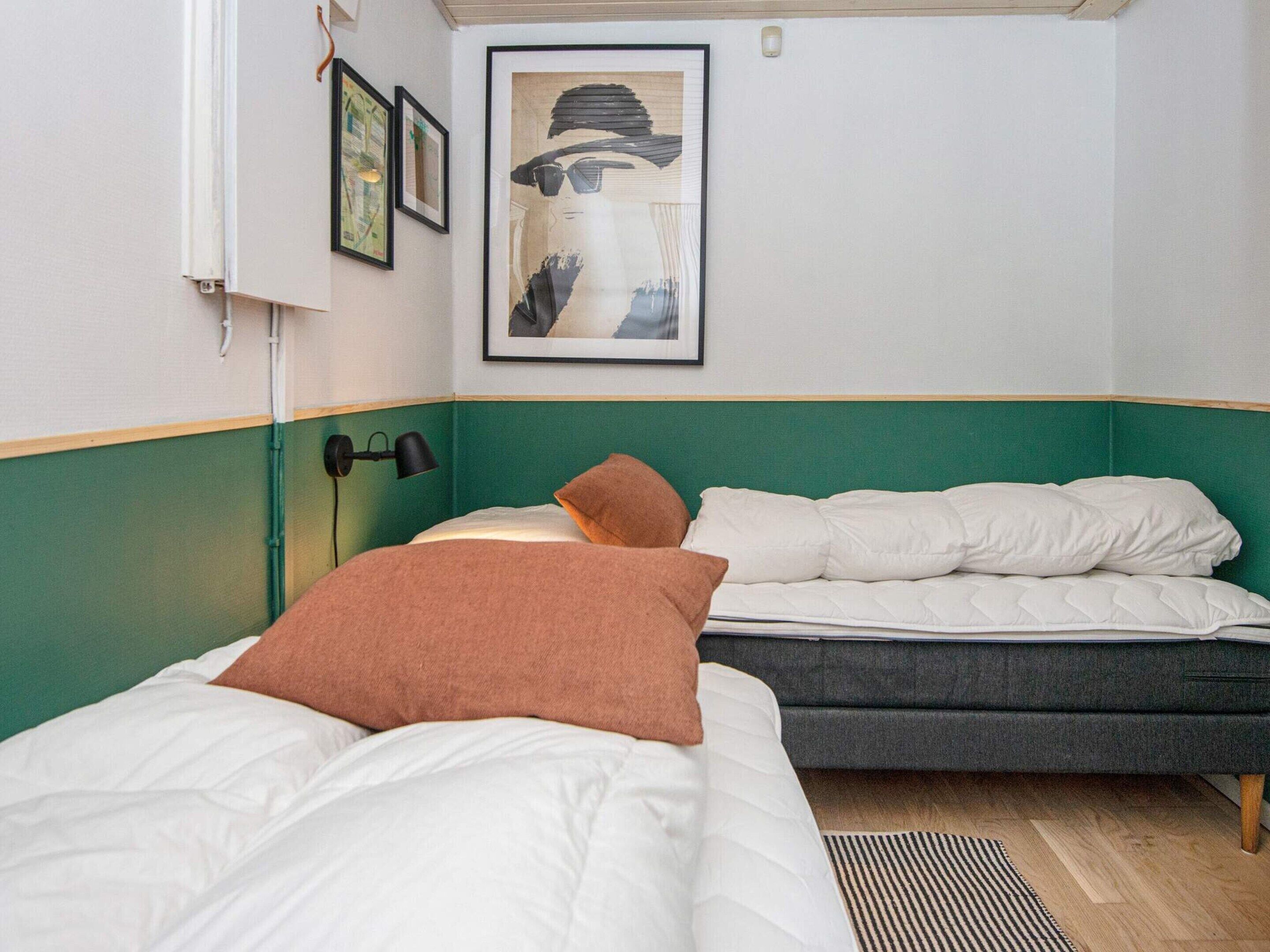 A room with two beds, one with a brown pillow and the other with white pillows. There is a framed picture on the wall and a lamp on the side.