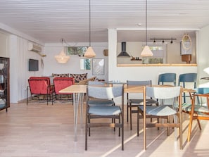 Un grand espace de salle à manger avec plusieurs tables et chaises, un coin cuisine avec des luminaires suspendus et un coin salon avec un fauteuil rouge.