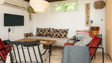 A cozy living room with a wooden table, striped chairs, a sofa with patterned cushions, a television, and a window with a view of greenery.