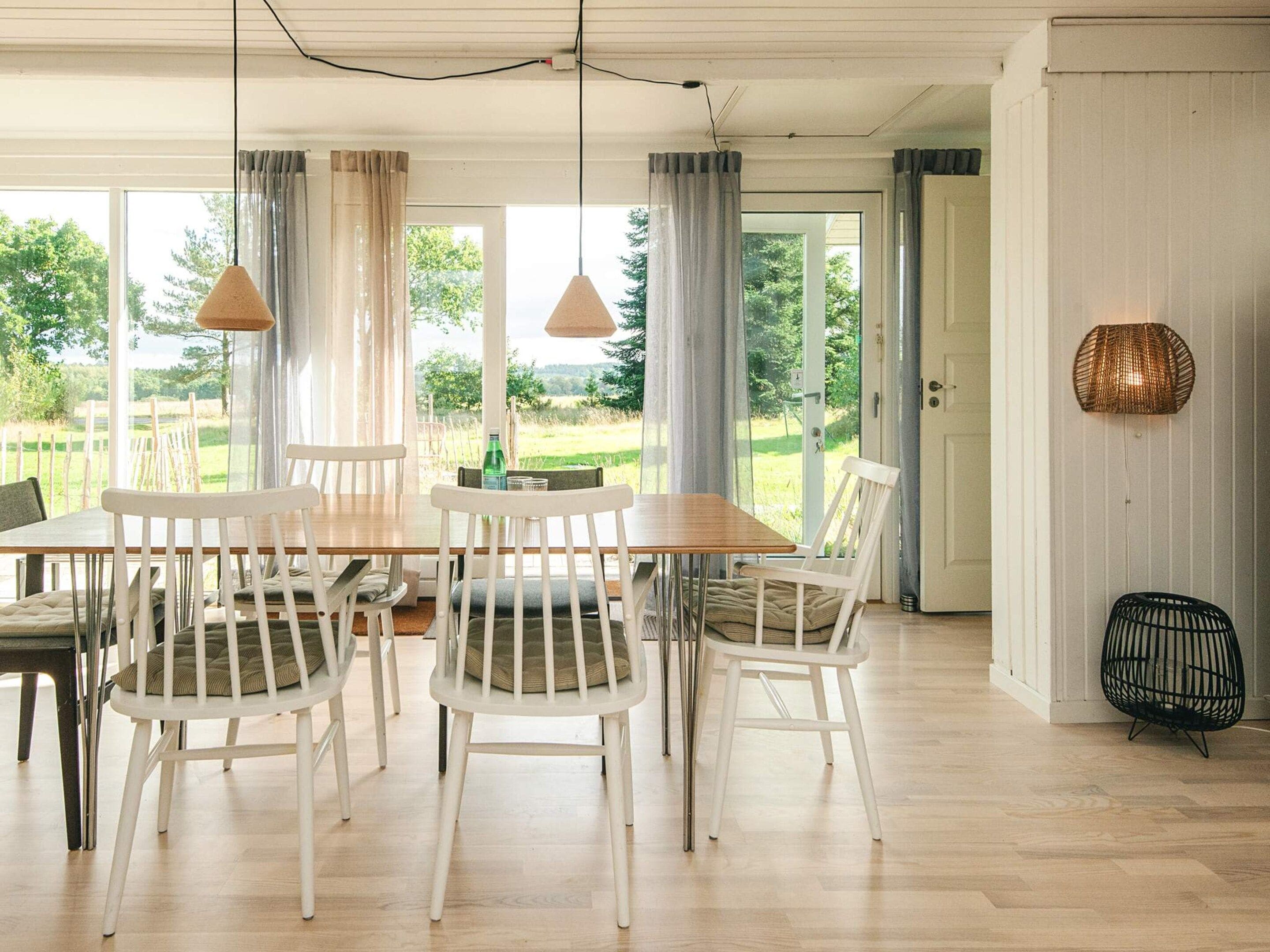 A dining area with a wooden table and white chairs, large windows with curtains, and a view of greenery outside.