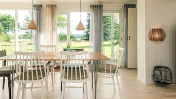 A dining area with a wooden table and white chairs, large windows with curtains, and a view of greenery outside.