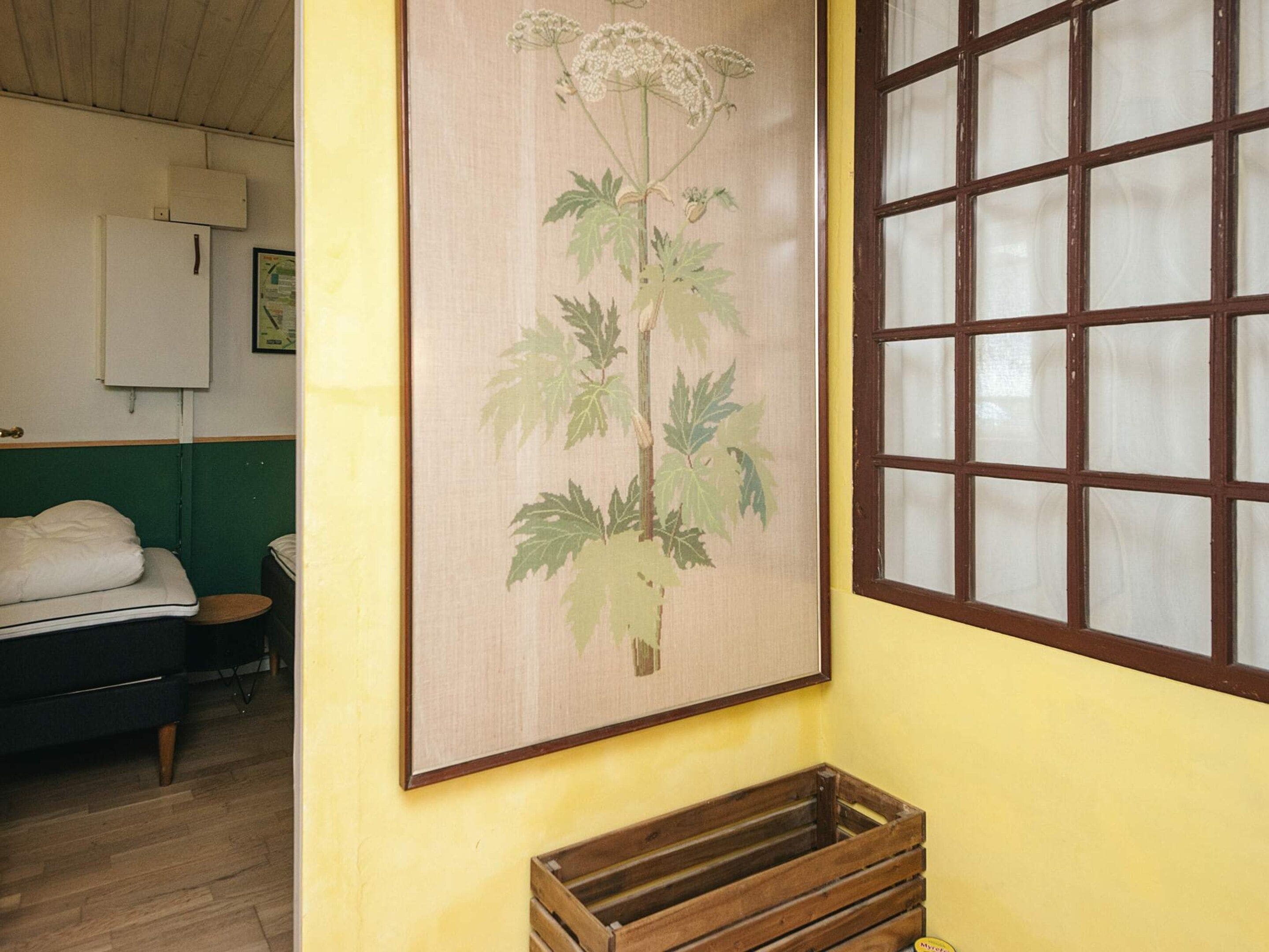 A room with a bed, a wooden crate, a framed botanical illustration, and a window with a grid pattern.