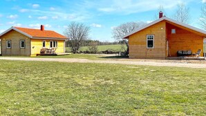 Two wooden cabins with attached porches, a dirt path between them, and a grassy field in the background.