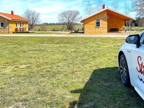 A white car parked on a grassy field with two wooden buildings in the background.
