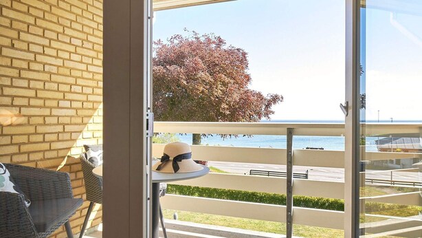 Ein Balkon mit Meerblick, ein Korbstuhl, ein kleiner runder Tisch mit einem Hut und eine Ziegelwand.