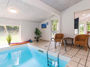 An indoor swimming pool with a ladder, wicker chairs, a small table, and a potted plant.