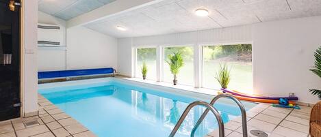 An indoor swimming pool with tiled flooring, a blue pool edge, and metal handrails.