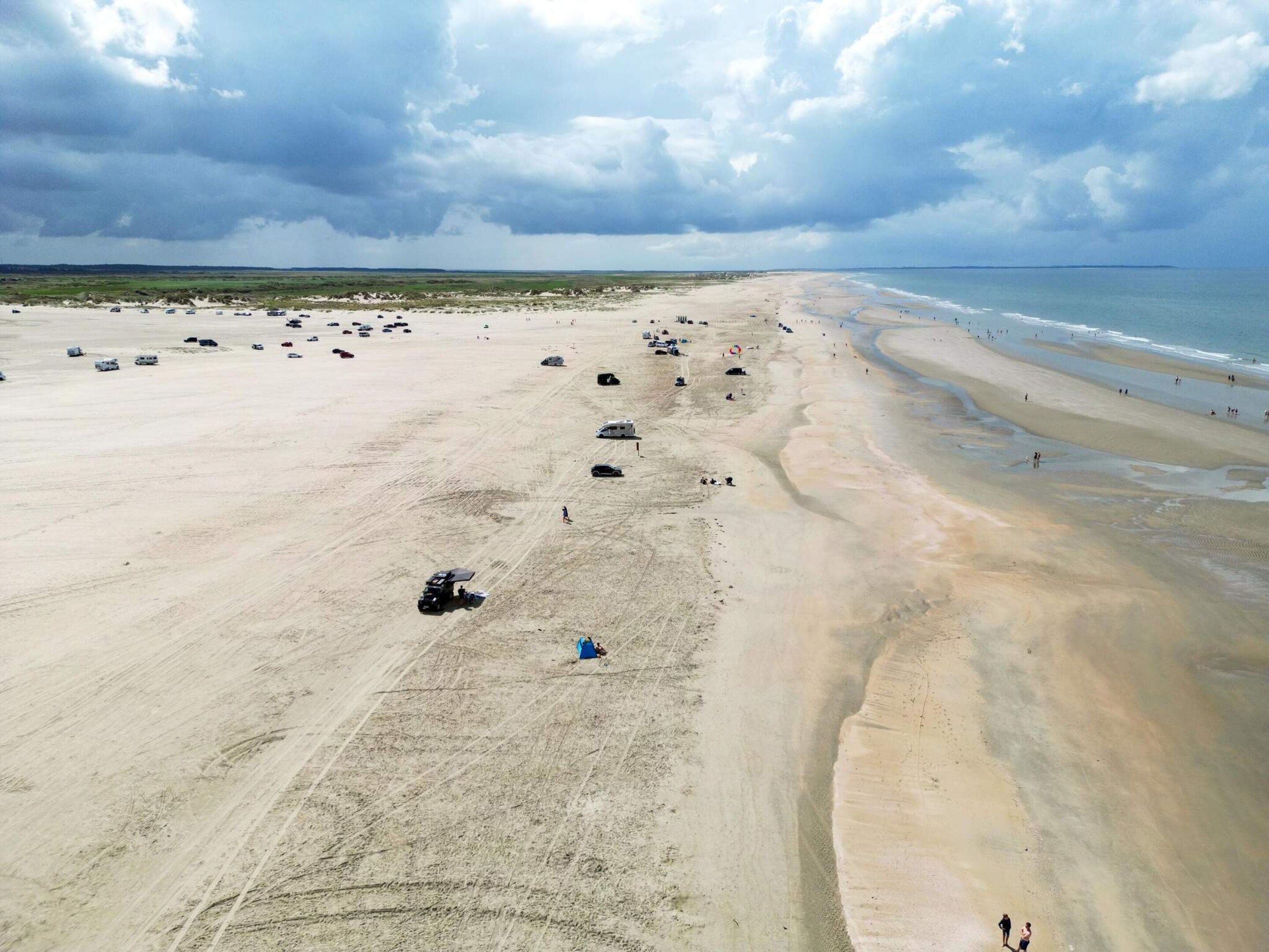 Uma praia extensa com veículos estacionados, céu claro e uma linha costeira distante.