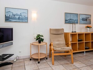 A living room with a television, a wooden chair, a small table with a potted plant, and a wooden shelf with decorative items. - 8 Person Holiday Home in Romo (Rømø)