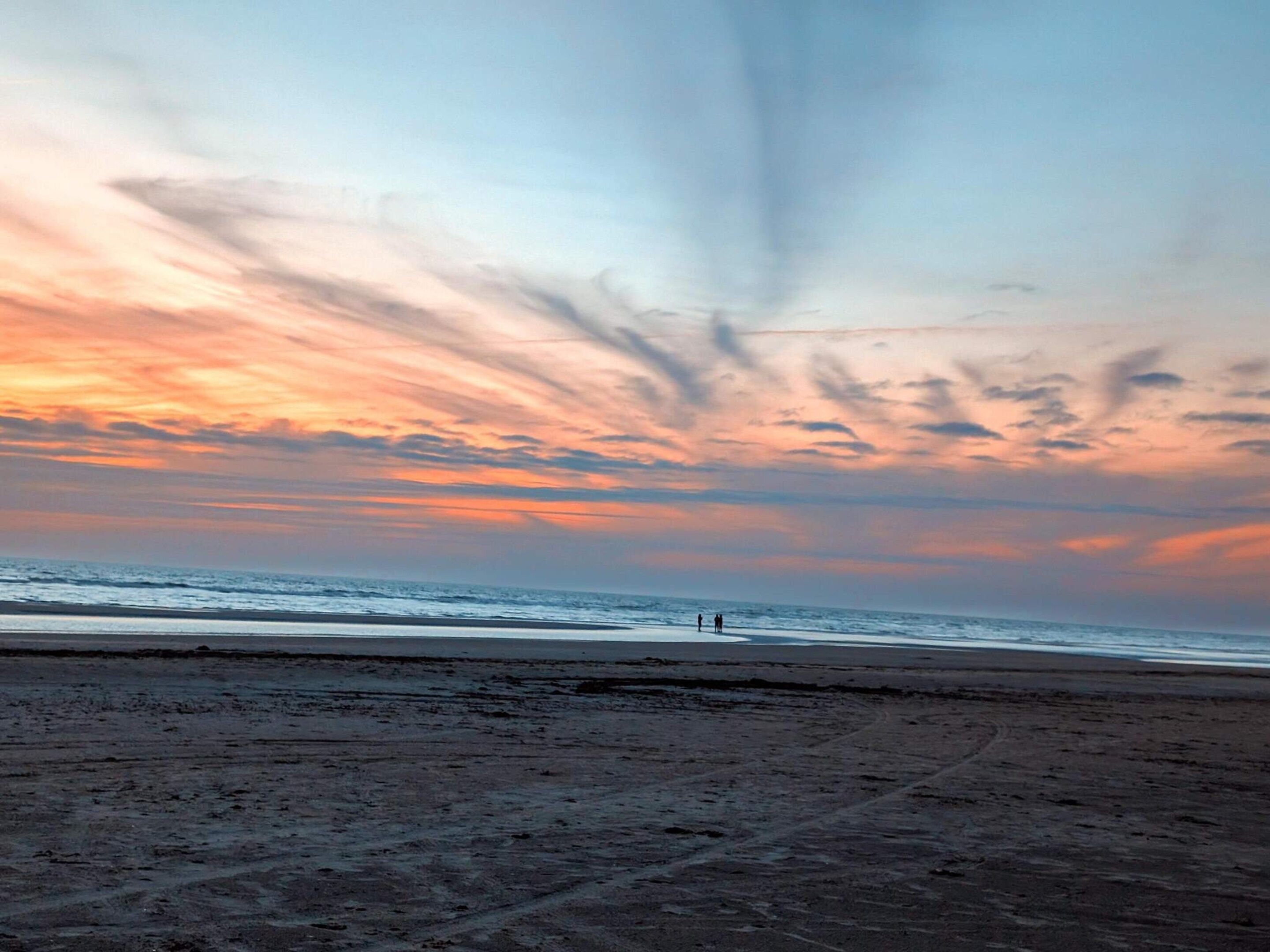 Una playa con cielo al atardecer, terreno arenoso y dos personas a lo lejos.