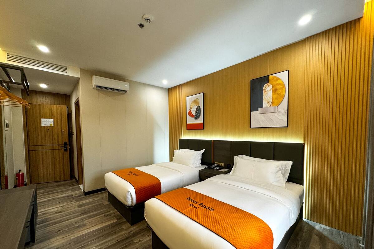 A hotel room with two beds, wooden flooring, a wooden door, an air conditioning unit, and two framed pictures on the wall.