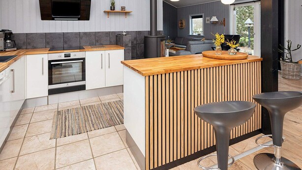 A modern kitchen with a wooden island, built-in oven, and a flat-screen TV. There are two bar stools and a small seating area with a sofa and a coffee table. - 4 Person Holiday Home in Vinderup (Vinderup)