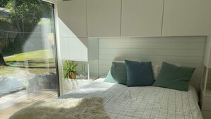 Ein Schlafzimmer mit einem großen Bett, einem weißen Kopfteil und Blick auf einen Garten durch eine Schiebetür.