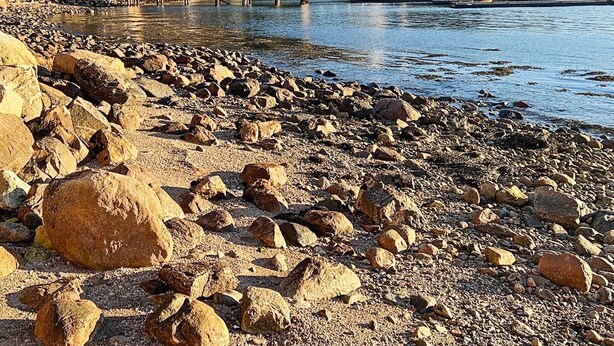 Una costa rocosa con un muelle de madera que se adentra en aguas tranquilas, rodeada de colinas y vegetación escasa.