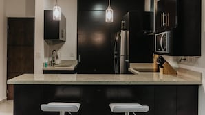 Une cuisine moderne avec des meubles de couleur sombre, un îlot central doté de tabourets de bar, des appareils en acier inoxydable et un éclairage suspendu.
