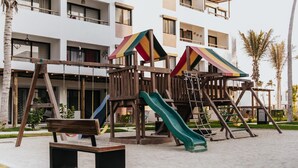 Un terrain de jeux avec un toboggan et des balançoires, situé devant un immeuble doté de balcons et de fenêtres.