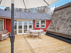 A wooden deck with a table and chairs, a thatched roof structure, and a red building with large windows. - 4 Star Holiday Home in Aakirkeby (Akirkeby)