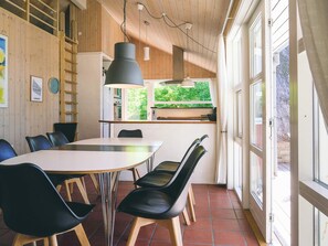 A dining area with a round table and chairs, wooden walls, a window with a view of greenery, and a kitchen area with cabinets and a stove.