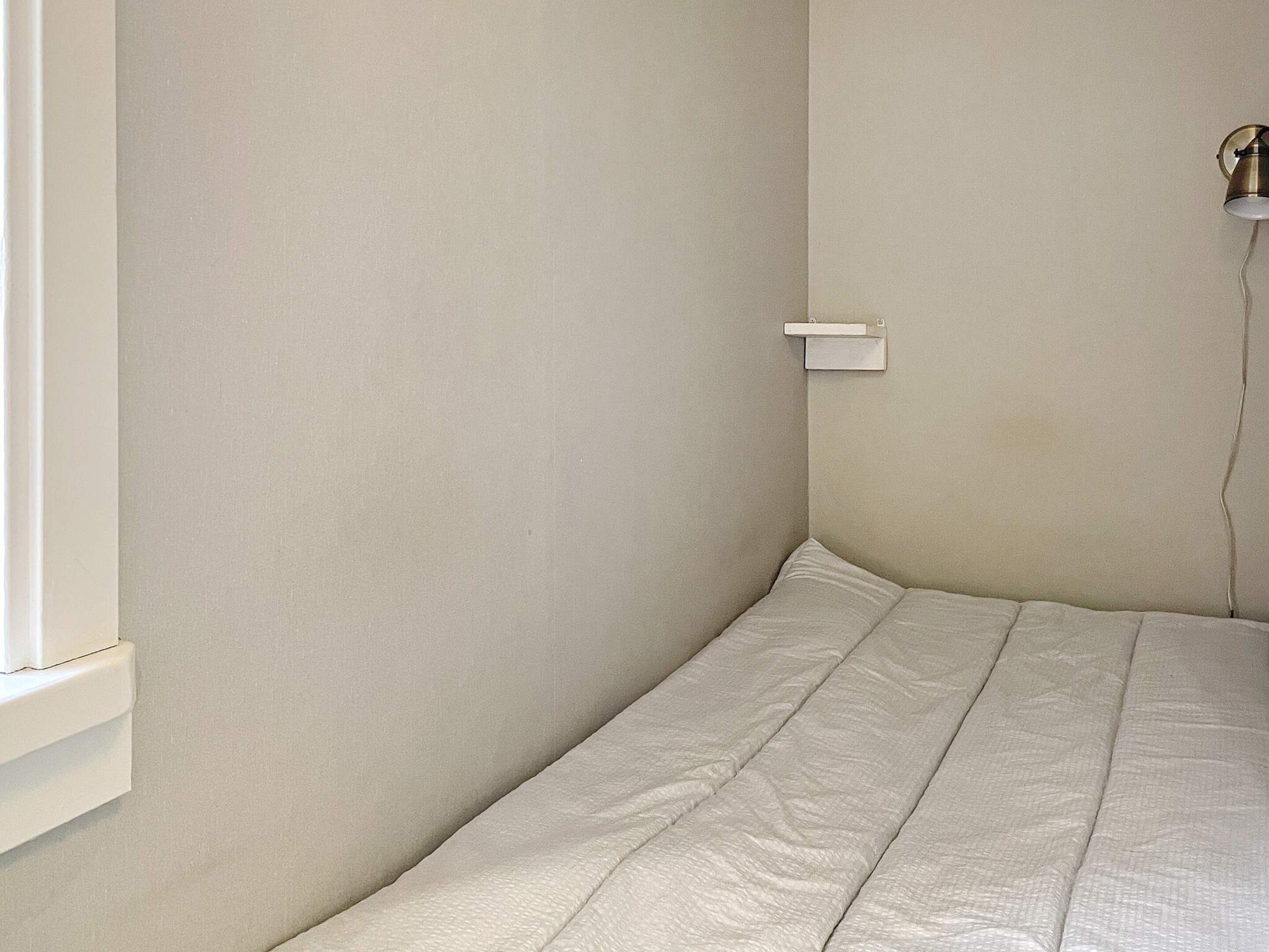 A small, single-bed room with a plain wall and a window on one side.