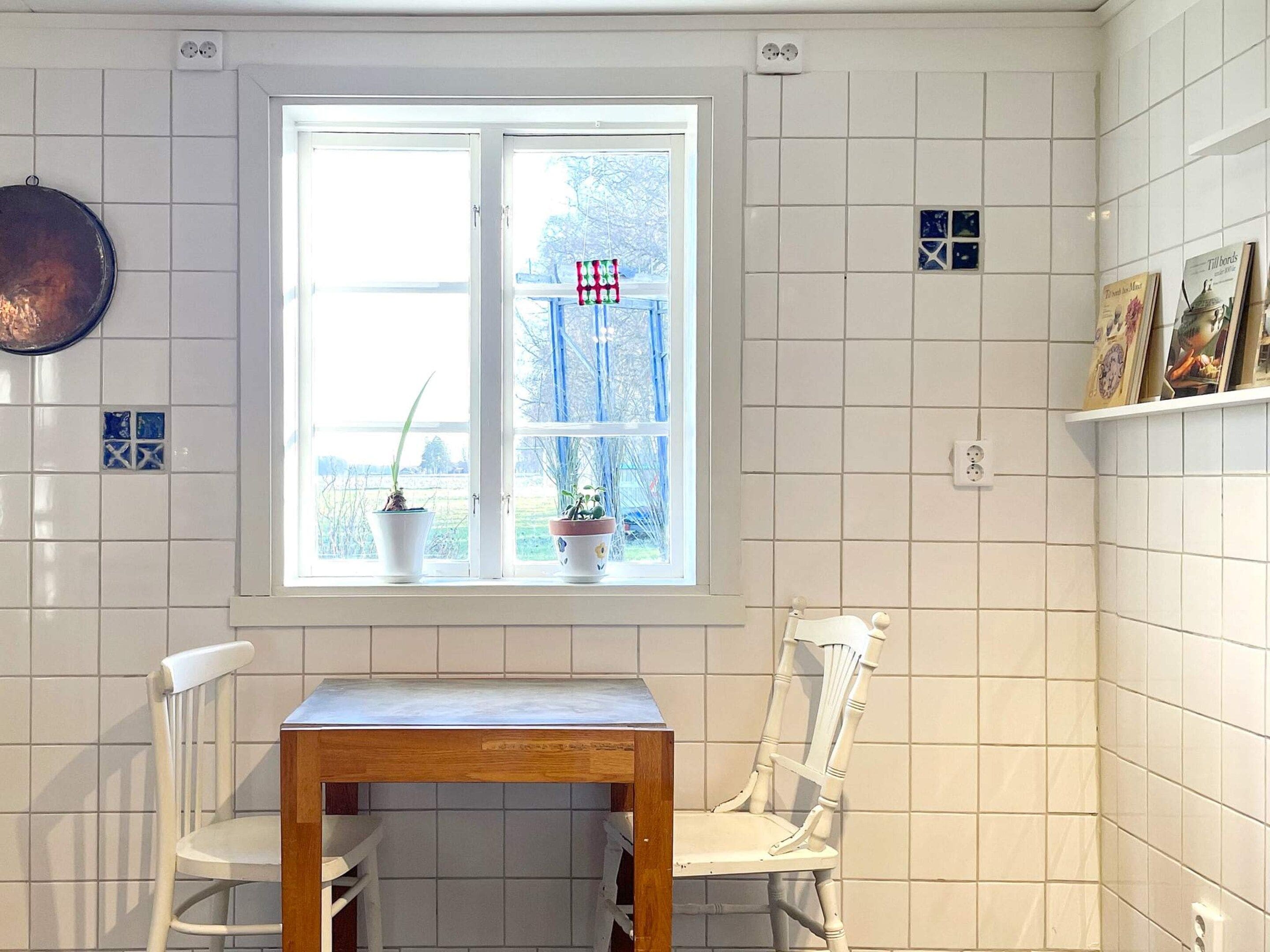 A kitchen with white tiled walls, a window with two potted plants, a wooden table, and two white chairs.