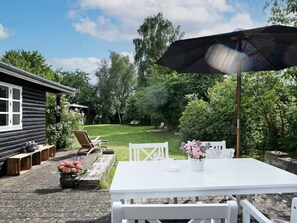 A patio with a white table and chairs, a wooden bench, and a garden with a lawn and trees. - 4 Person Holiday Home in Helsinge (Helsinge)