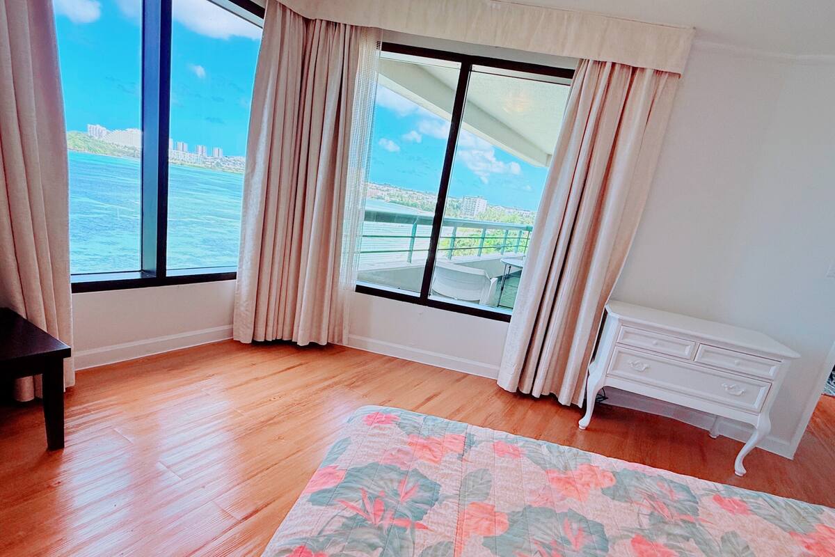 Una habitación con un amplio ventanal que ofrece vistas al mar y edificios, suelo de madera, alfombra estampada y cómoda blanca.