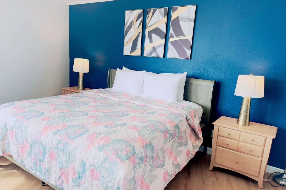 Un dormitorio con una cama, dos mesitas de noche, una mesita auxiliar, una pared azul y tres cuadros abstractos.