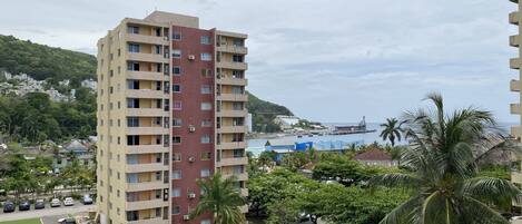 A high-rise building with multiple balconies, a parking area with cars, and a view of the sea and a dock.