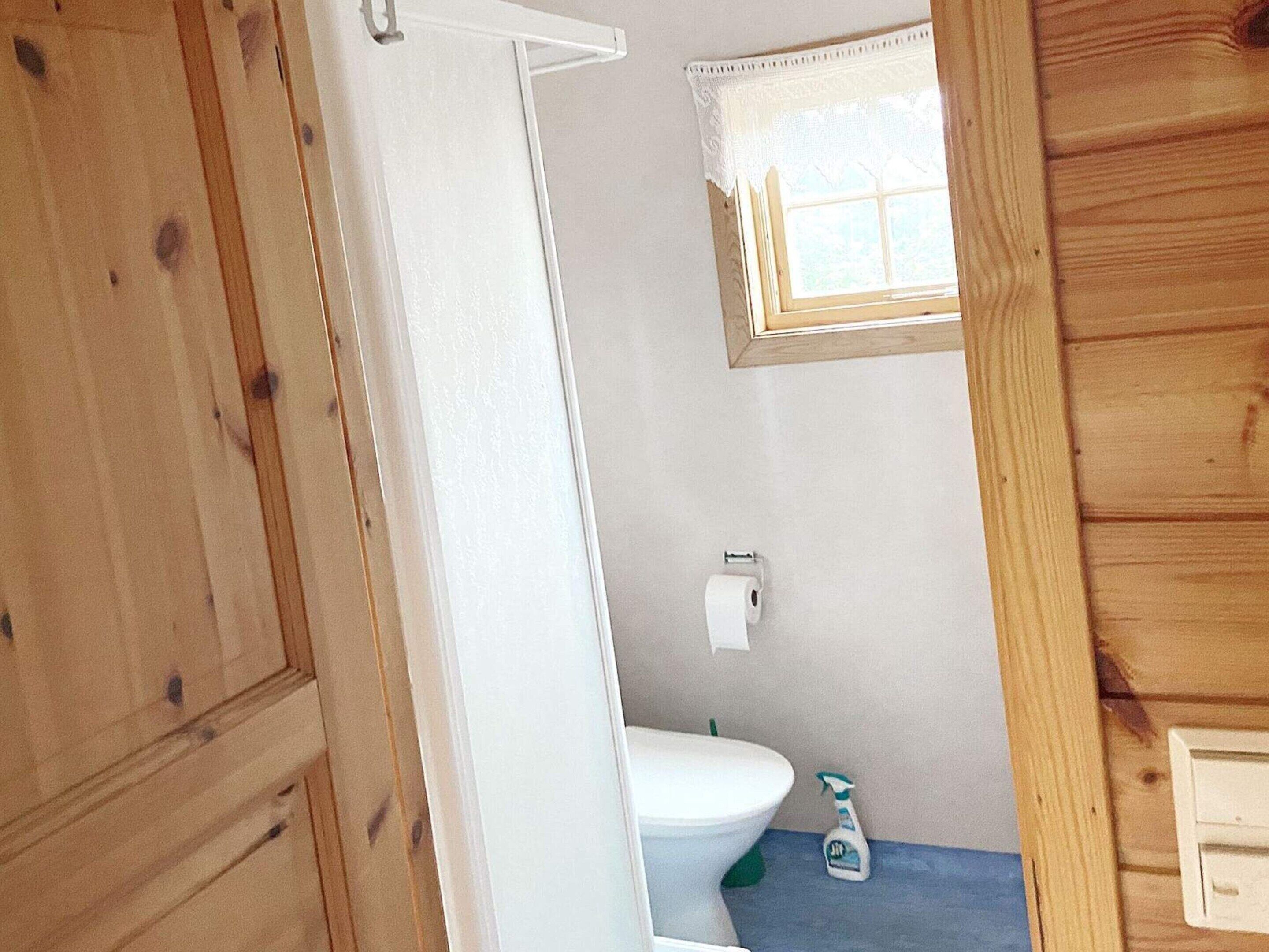 A wooden cabin bathroom with a toilet, a small window with lace curtains, and a wooden cabinet.