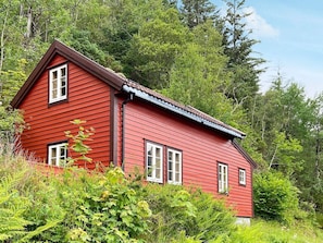Une maison en bois rouge, dotée d’un toit à pignon et entourée d’un feuillage verdoyant et luxuriant.