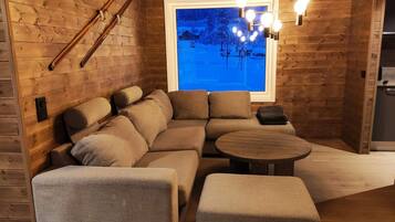 A cozy wooden interior with a sectional sofa, a round coffee table, and a window with a view of a snowy landscape.