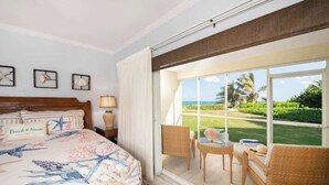 Habitación de hotel con cama, ventana con vista al océano y follaje, pequeña mesa con sillas y una lámpara.