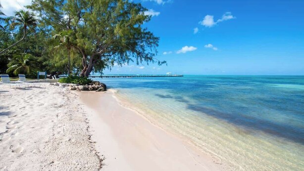 Una playa con agua turquesa cristalina, una orilla arenosa y un muelle a lo lejos.