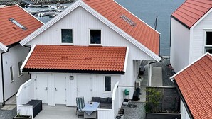 Ein weißes Haus mit rotem Ziegeldach, einer Terrasse mit Tisch und Stühlen und Blick auf einen See mit Booten und Berge im Hintergrund.