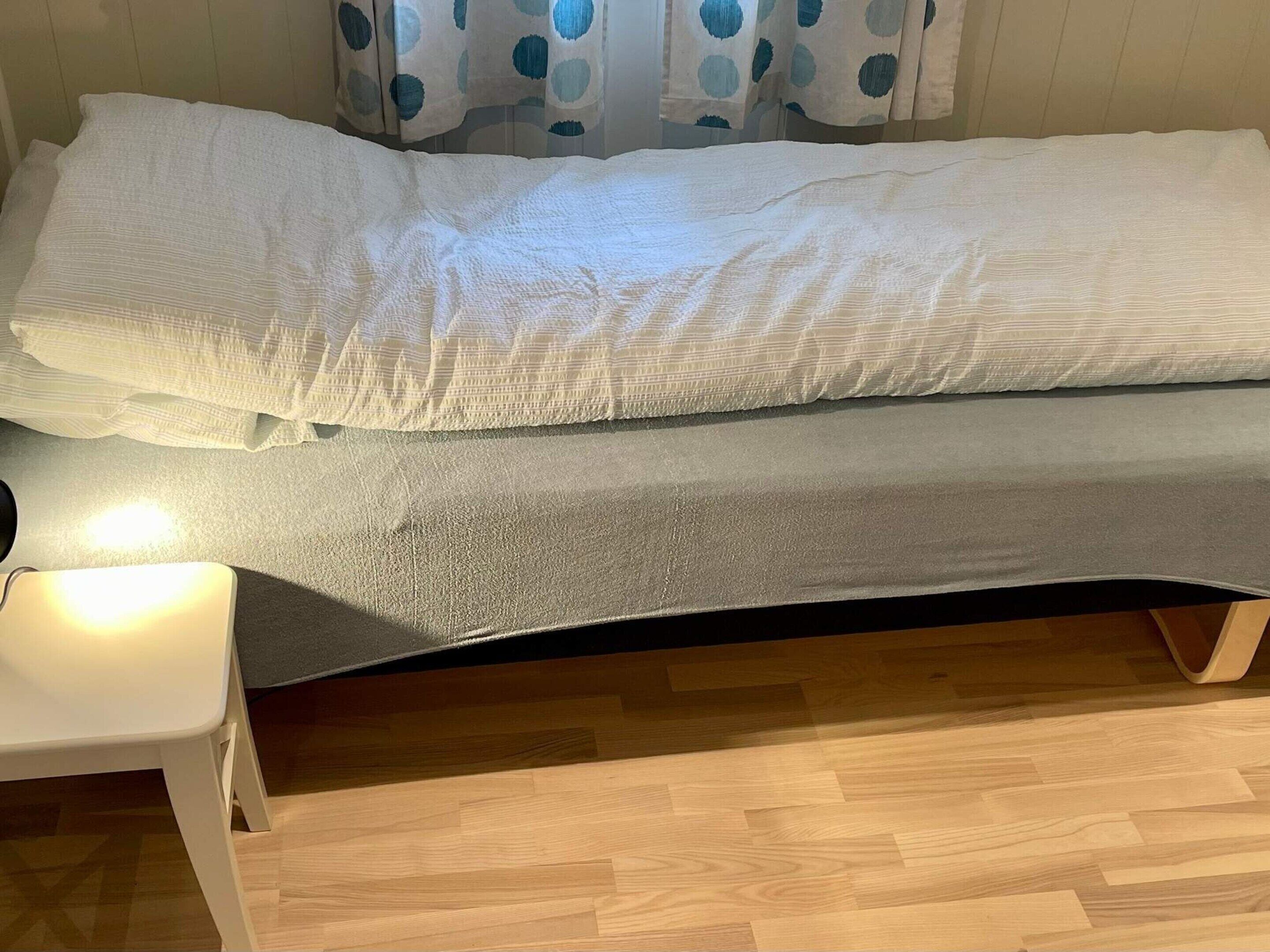 A single bed with a grey mattress and white pillows, a bedside table, and a wooden floor.