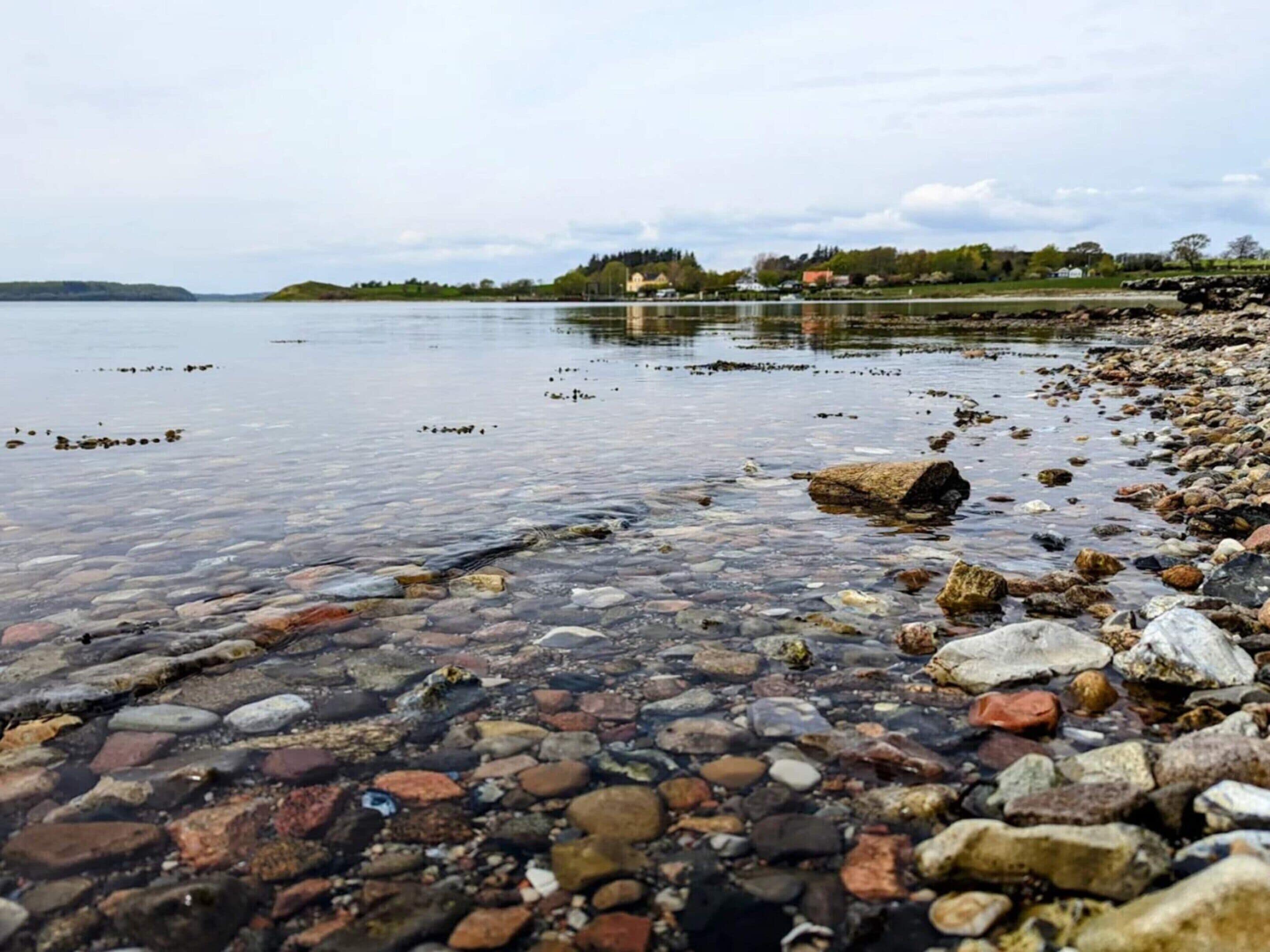 A pebbly shoreline with clear water, a distant building, and a calm lake.