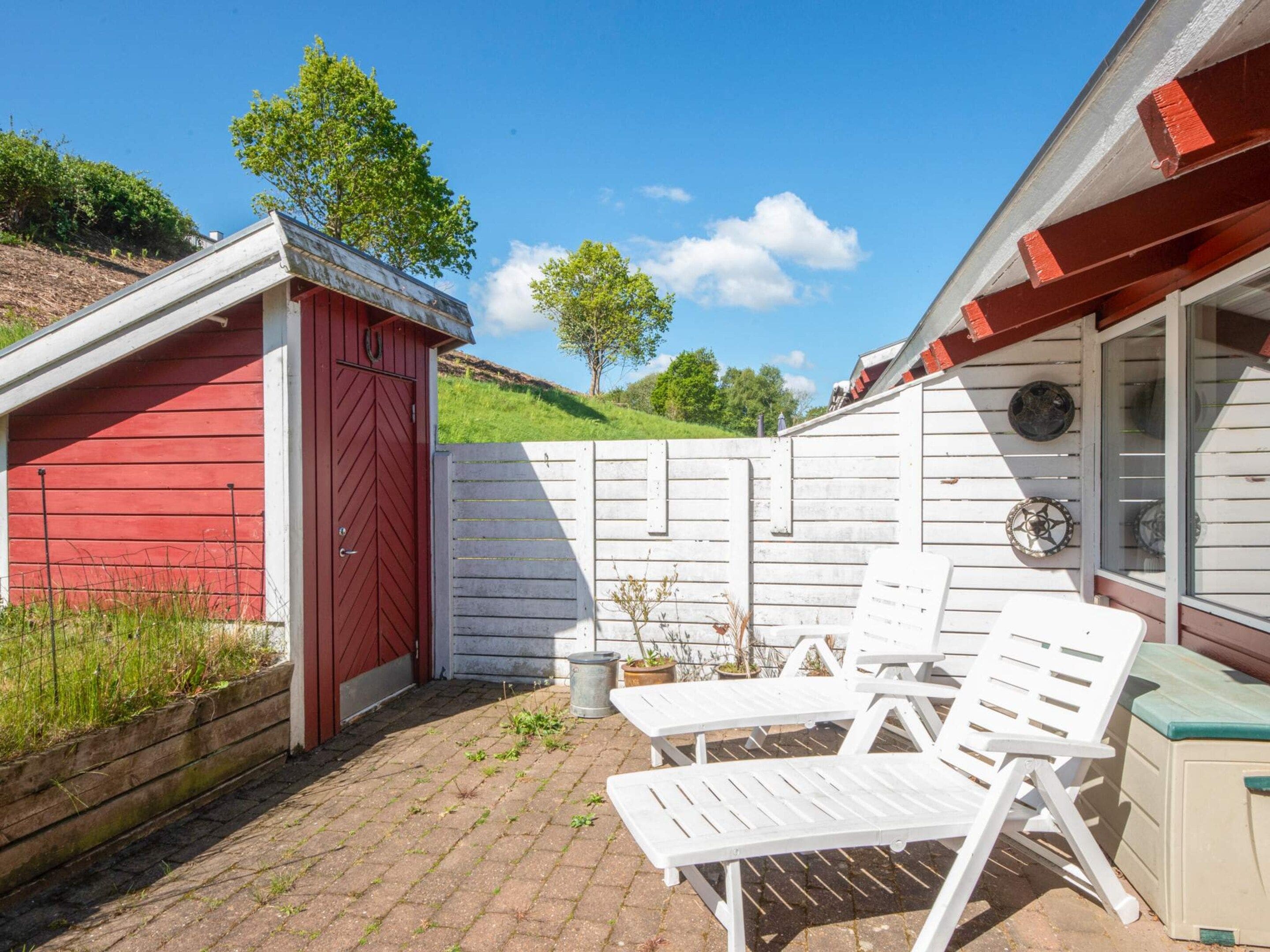 Eine Terrasse mit weißen Plastikstühlen und einem Tisch, ein rotes Gartenhaus und ein weißer Zaun.