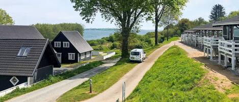 A rural setting with a dirt path, a white van, and a row of wooden cabins.