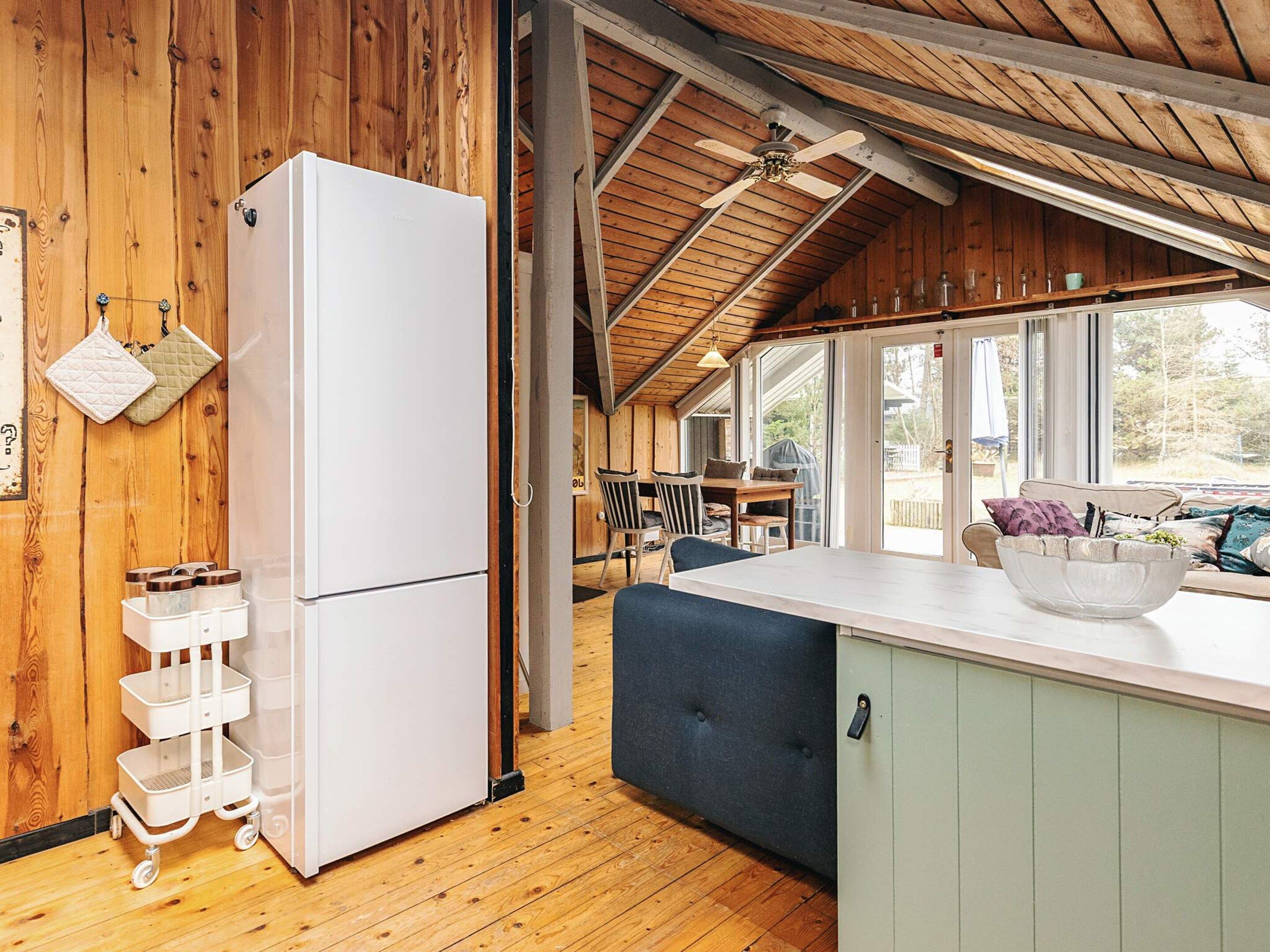 Un intérieur en bois, avec un réfrigérateur blanc, un coin repas comprenant des chaises et une table, et une grande fenêtre.