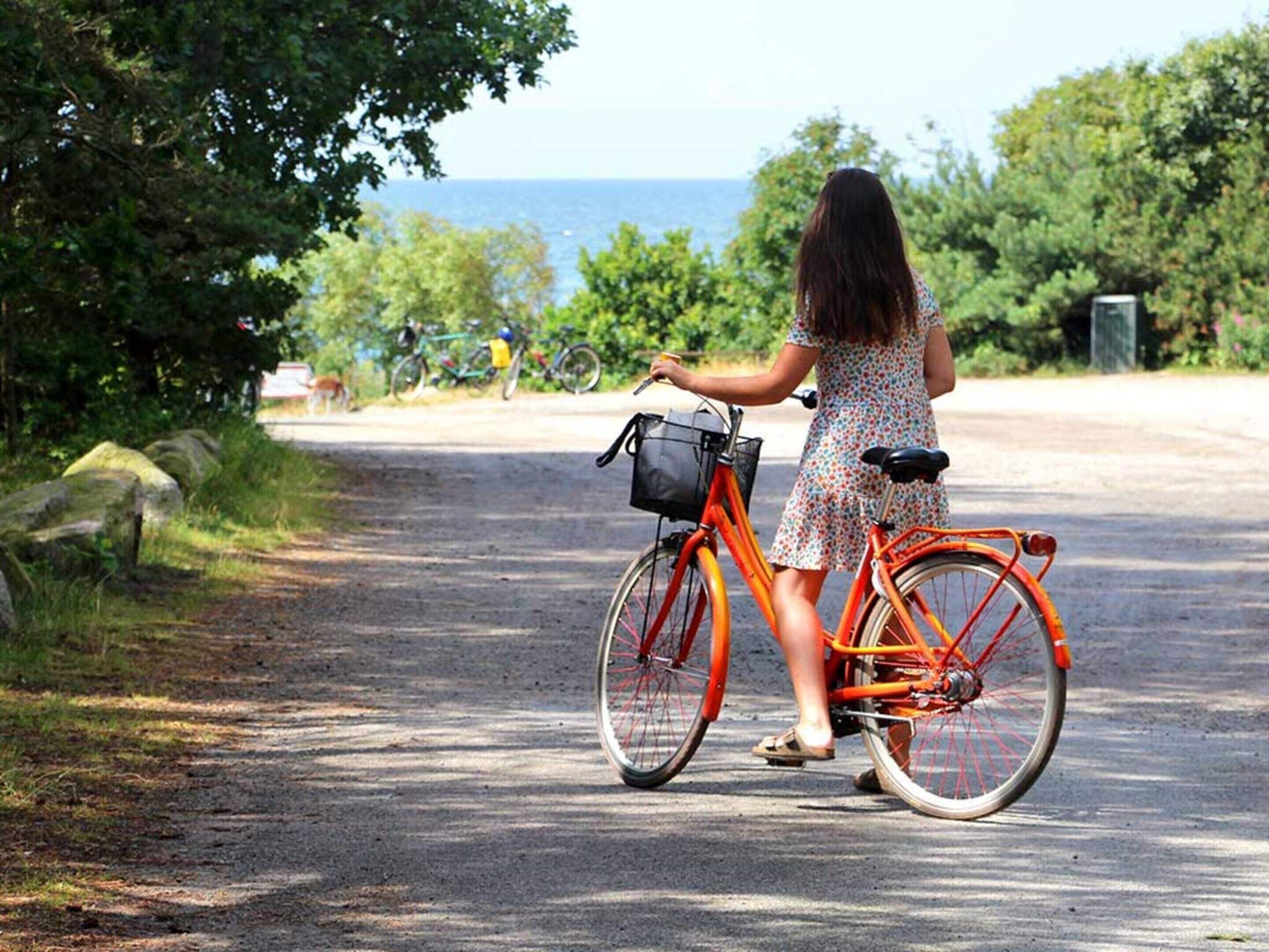 Eine Frau steht neben einem orangefarbenen Fahrrad mit Korb, auf einem befestigten Weg mit Grünflächen und Blick aufs Meer im Hintergrund.