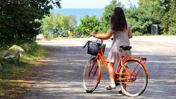 A woman standing next to an orange bicycle with a basket, on a paved path with greenery and a view of the sea in the background.