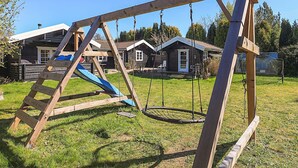 En gungställning i trä på en gräsig gård, med en rutschkana, omgiven av trähus.