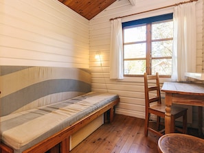 Ein Blockhauszimmer mit einer Holzbänkl, einem Tisch und einem Vorhangfenster.