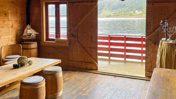 Interior de una cabaña de madera con una mesa de comedor, taburetes, un pequeño modelo de barco y vistas de un lago a través de un ventanal amplio.