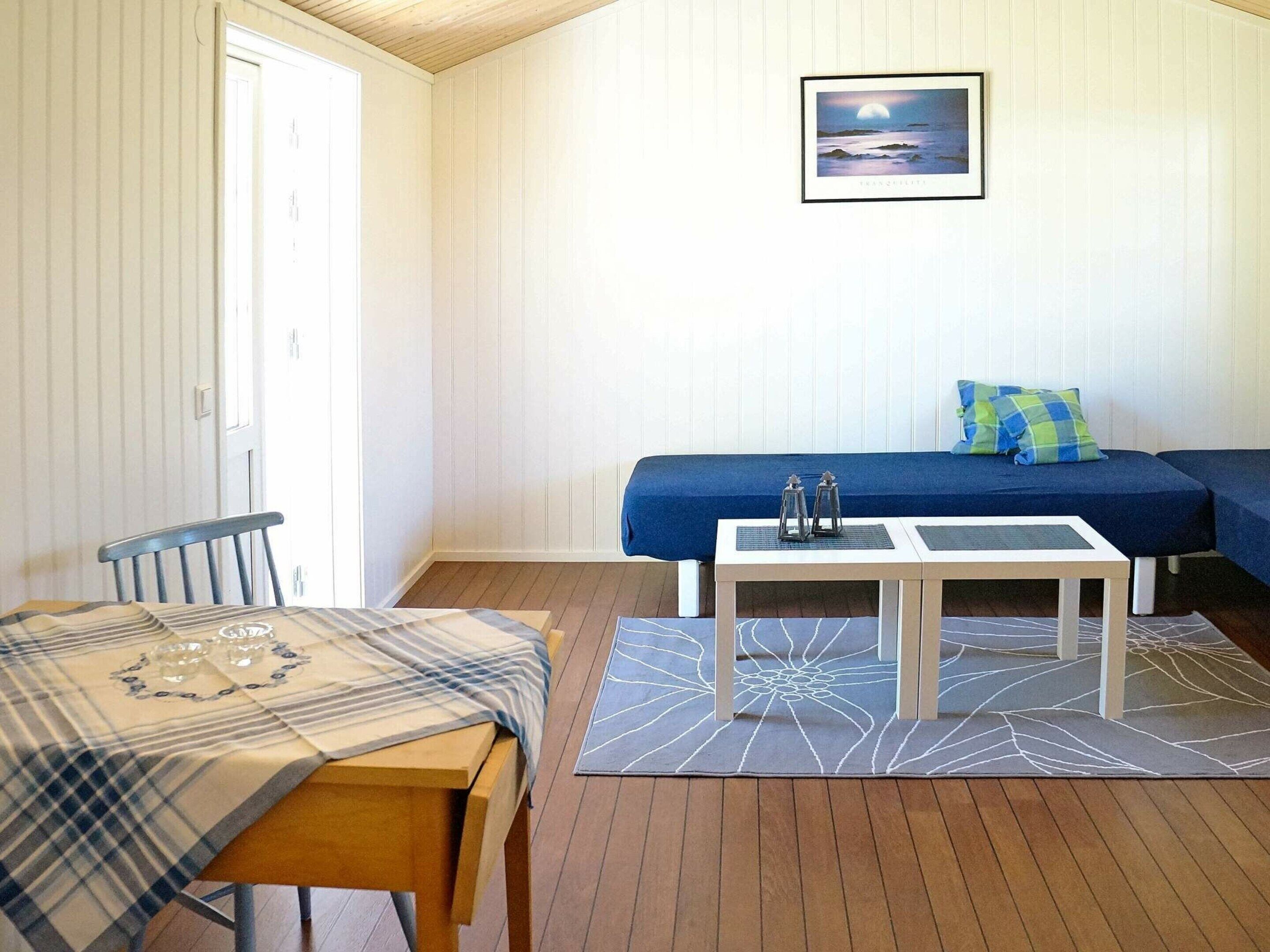 Ein Zimmer mit Holzboden, einem Bett mit blauer Bettwäsche, einem Tisch mit kariertem Tischläufer, einem Stuhl und einem gerahmten Bild an der Wand.