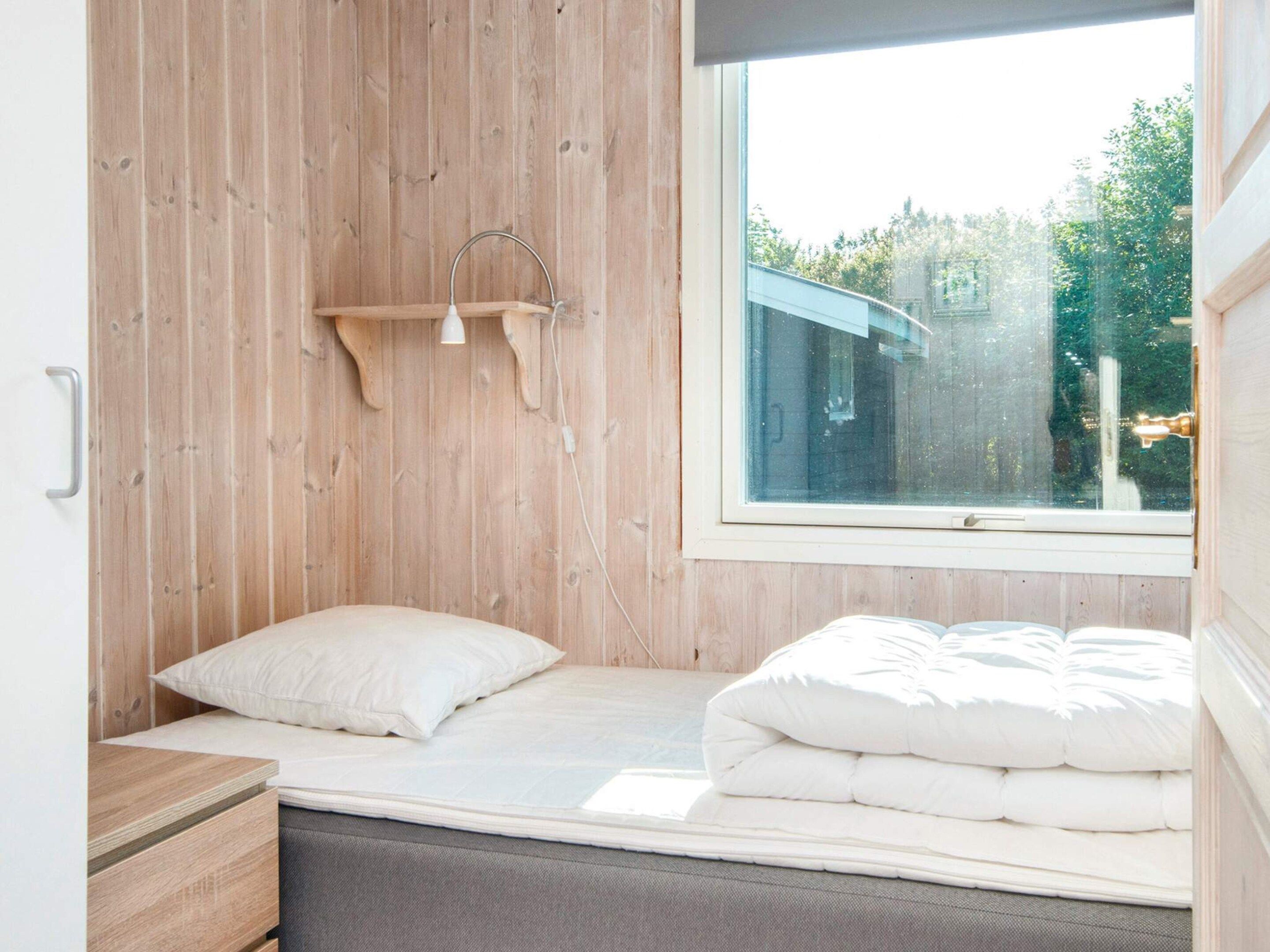 A small, single-bed room with a wooden wall, a window, and a bedside table.